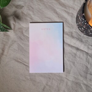 Könnte beinhalten: Ein Notizblock mit einem pastellfarbenen Aquarelldesign in Rosa, Blau und Weiß. Das Wort "NOTES" ist in hellrosa Schrift oben aufgedruckt. Der Notizblock liegt auf einer beigen Leinenoberfläche.