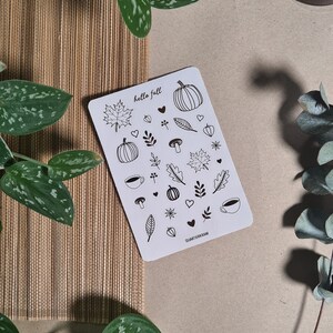 Può includere: Una carta bianca con illustrazioni a tratto nero a tema autunnale, tra cui zucche, foglie, cuori e tazze di caffè. Le parole "hello fall" sono scritte in alto. La carta è circondata da piante verdi e un tappetino intrecciato marrone.