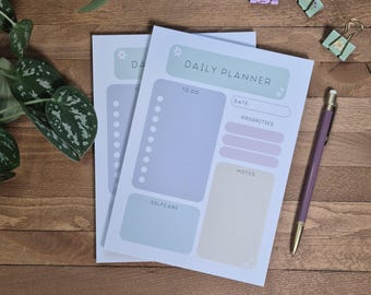 Op de afbeelding: Twee pastelblauwe en paarse dagelijkse plannerpagina's met bloemmotieven. De bovenste pagina heeft de tekst "DAILY PLANNER" en secties voor "TO DO", "DATE", "PRIORITIES", "NOTES" en "SELFCARE".