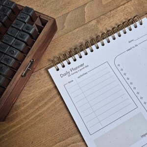 Może przedstawiać: Biały planer dzienny ze złotą spiralą. Planer zawiera sekcje: "time", "activities", "need to do" i "want to do". Na górze strony widnieje napis "Daily Planner".