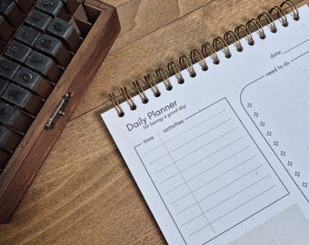 May include: A white daily planner with a gold spiral binding. The planner has a section for "time", "activities", "need to do", and "want to do". The text "Daily Planner" is printed at the top of the page.