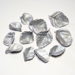 May include: A collection of silver-colored metal leaves, each with detailed veining, scattered on a white surface. The leaves vary in size and shape, resembling natural foliage.