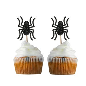 Puede incluir: Dos cupcakes con adornos de cupcake de araña de purpurina negra. Los cupcakes están cubiertos con glaseado blanco y tienen una base marrón claro.