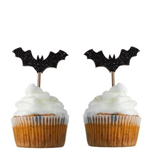 Puede incluir: Dos cupcakes con glaseado blanco y adornos de murciélago negro brillante. Los cupcakes tienen envoltorios de papel marrón y blanco. Los murciélagos están sobre palitos de madera. Fondo blanco.