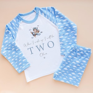 May include: A two-piece pyjama set with a white top and blue sleeves, plus matching blue trousers. The top features a bear in an aeroplane graphic and the text "When I wake up I will be TWO Oliver". The sleeves and trousers have a cloud pattern.