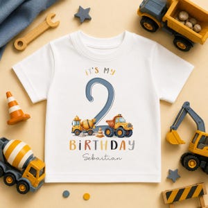 May include: A white short-sleeve t-shirt featuring a large blue number 2, yellow construction trucks, and the text "it's my BIRTHDAY" with the name "Sebastian" printed below. The graphic includes a cement mixer and a dump truck.