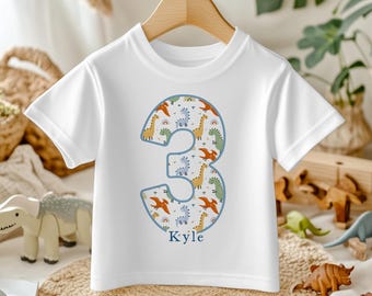 May include: White short-sleeve t-shirt featuring a large number "3" filled with a dinosaur pattern in orange, green, and yellow. The name "Kyle" is printed below the number. The shirt is hanging on a wooden hanger.