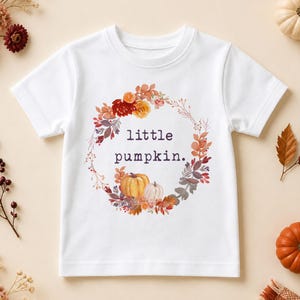 May include: A white short-sleeved t-shirt featuring a central graphic of a circular autumn-themed wreath made of orange, red, and yellow leaves and flowers. Inside the wreath, the text reads little pumpkin in a dark purple serif font.
