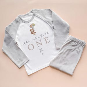 May include: A baby's light grey and white pajama set. The long-sleeved top has grey sleeves and a white front with a teddy bear graphic holding balloons. Text on the front reads "When I wake up I will be ONE" and the name "Theo".