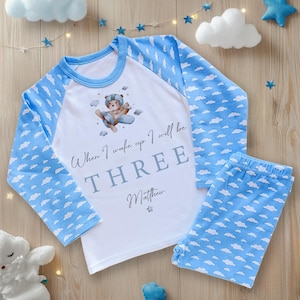 May include: A child's two-piece pyjama set. The top is white with blue sleeves patterned with white clouds. The front of the top has a graphic of a bear in a plane and the text "When I wake up I will be THREE Matthew". The bottoms are blue with white clouds.
