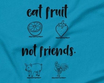 Puede incluir: Una camiseta azul turquesa con el texto "eat fruit not friends." El texto está en negro y hay ilustraciones de un limón, una fresa, un cerdo y un gallo.