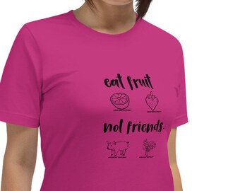 Puede incluir: Una camiseta rosa con el texto "eat fruit not friends." El texto está en negro y hay pequeñas ilustraciones negras de una fresa, una naranja, un cerdo y un pollo.