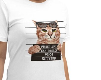 May include: White t-shirt with a humorous design of a cat in sunglasses, styled as a police mugshot. The cat is brown and tan, with the text "POLICE DEPT. SAN DIEGO CA 92104 KITTY BAD" on a sign.