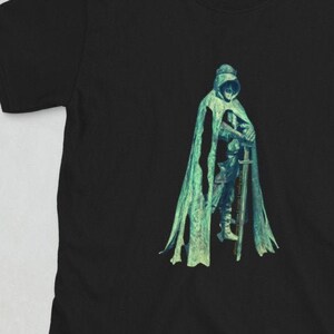 May include: Black t-shirt featuring a graphic of a hooded figure in a green cloak holding a sword.