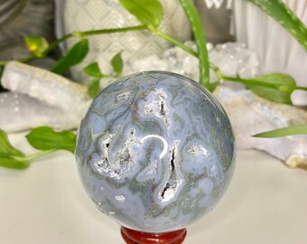 May include: A polished spherical stone featuring a complex pattern of muted blue and grey tones with intricate, web-like veining and small, crystalline vugs. The sphere rests securely on a small, circular, dark reddish-brown wooden display stand.