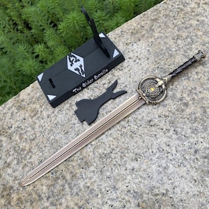 Pu&ograve; includere: Una spada decorativa con una lama in bronzo e un'elsa ornata, accompagnata da un supporto nero e un pezzo nero separato. Il supporto presenta la scritta "The Elder Scrolls" e un simbolo. La lama della spada ha un design a strisce.