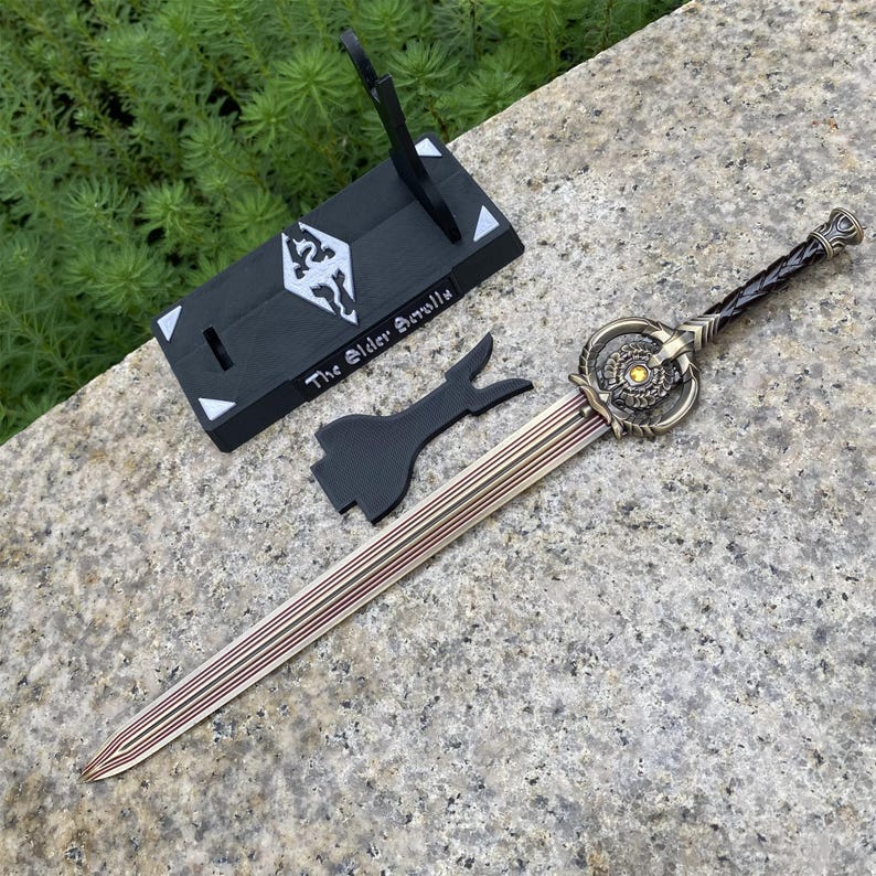 Pu&ograve; includere: Una spada decorativa con una lama in bronzo e un'elsa ornata, accompagnata da un supporto nero e un pezzo nero separato. Il supporto presenta la scritta "The Elder Scrolls" e un simbolo. La lama della spada ha un design a strisce.