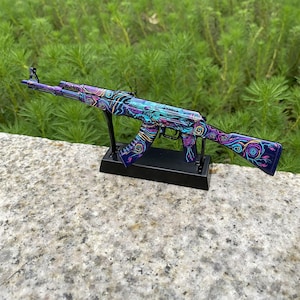 May include: A decorative AK-47 rifle model with a vibrant, swirling design in shades of purple, blue, and teal. The model is mounted on a black stand, showcasing the intricate patterns. The background features lush green foliage.