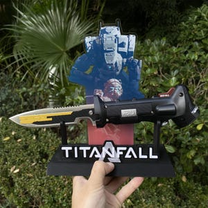 Może przedstawiać: Czarna i złota replika noża Titanfall ze stojakiem z napisem "TITANFALL". Nóż ma srebrne ostrze ze złotymi akcentami i czarną rękojeść. Za nożem znajduje się niebiesko-biała grafika robota.
