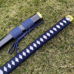 Może przedstawiać: Japoński miecz katana z czarną pochwą i granatowym uchwytem owiniętym we wz&oacute;r romb&oacute;w z białymi akcentami. Miecz spoczywa na zielonej trawie, prezentując tradycyjny design.