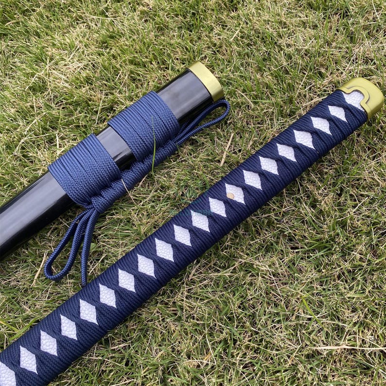 Może przedstawiać: Japoński miecz katana z czarną pochwą i granatowym uchwytem owiniętym we wz&oacute;r romb&oacute;w z białymi akcentami. Miecz spoczywa na zielonej trawie, prezentując tradycyjny design.