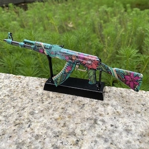 May include: A decorative AK-47 rifle model with a floral pattern in teal, pink, and green. The model is mounted on a black stand and set against a backdrop of green foliage. The rifle's design incorporates various flower and leaf motifs.