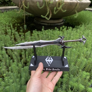 May include: A detailed replica of a sword from "The Elder Scrolls" video game series. The blade is silver with a black and red design. The sword rests on a black stand with the game's logo and text.
