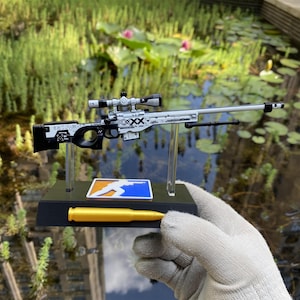 May include: A miniature white and black sniper rifle model with a scope, mounted on a black display stand. The stand features a rectangular blue and orange logo and a single gold-colored bullet replica attached to the front base section.