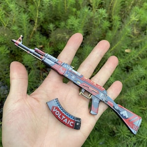 May include: A miniature AK-47 style rifle with a detached magazine. The rifle features a red, blue, and gray color scheme with intricate detailing. The magazine is black with the word "VOLTAIC" in red and white.