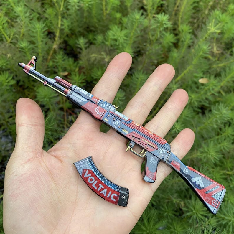 May include: A miniature AK-47 style rifle with a detached magazine. The rifle features a red, blue, and gray color scheme with intricate detailing. The magazine is black with the word "VOLTAIC" in red and white.