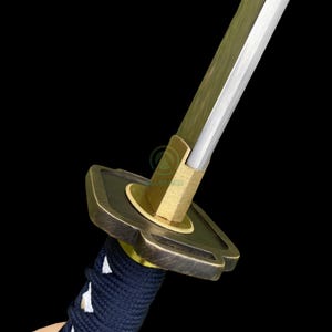 Może przedstawiać: Zbliżenie na miecz katana. Ostrze jest srebrne, a osłona jest brązowa z okrągłym wzorem. Rękojeść jest owinięta ciemnoniebieską tkaniną. Miecz jest umieszczony na czarnym tle.