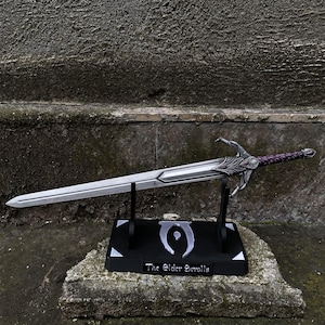 Könnte beinhalten: Ein silbernes Schwert mit dekorativem Griff und lila Griff liegt auf einem schwarzen Ständer. Der Ständer trägt den Schriftzug "The Elder Scrolls" und ein Symbol. Das Schwert wird auf einer Steinoberfläche präsentiert.