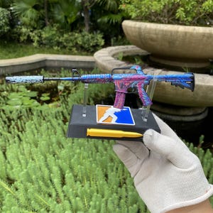 May include: A miniature rifle model featuring a vibrant blue and magenta marbled pattern, equipped with a black suppressor. The rifle is mounted on a black display stand with a square logo depicting a white silhouette and a yellow bullet-shaped accent.