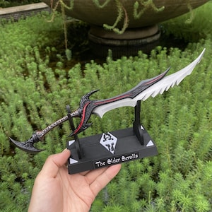 May include: A detailed replica of a fantasy sword, with a black and red handle and a silver blade with a serrated edge. The sword is displayed on a black stand with the text "The Elder Scrolls" and a logo.