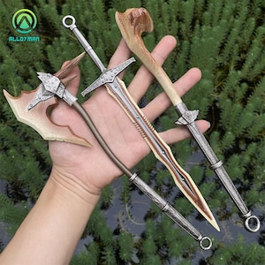 Puede incluir: Cuatro armas de fantas&iacute;a en miniatura: un hacha plateada con una hoja beige, una espada plateada con una hoja marr&oacute;n, una espada plateada con un mango de hueso y un garrote plateado con un mango de hueso. Las armas se sostienen sobre un fondo de follaje verde.