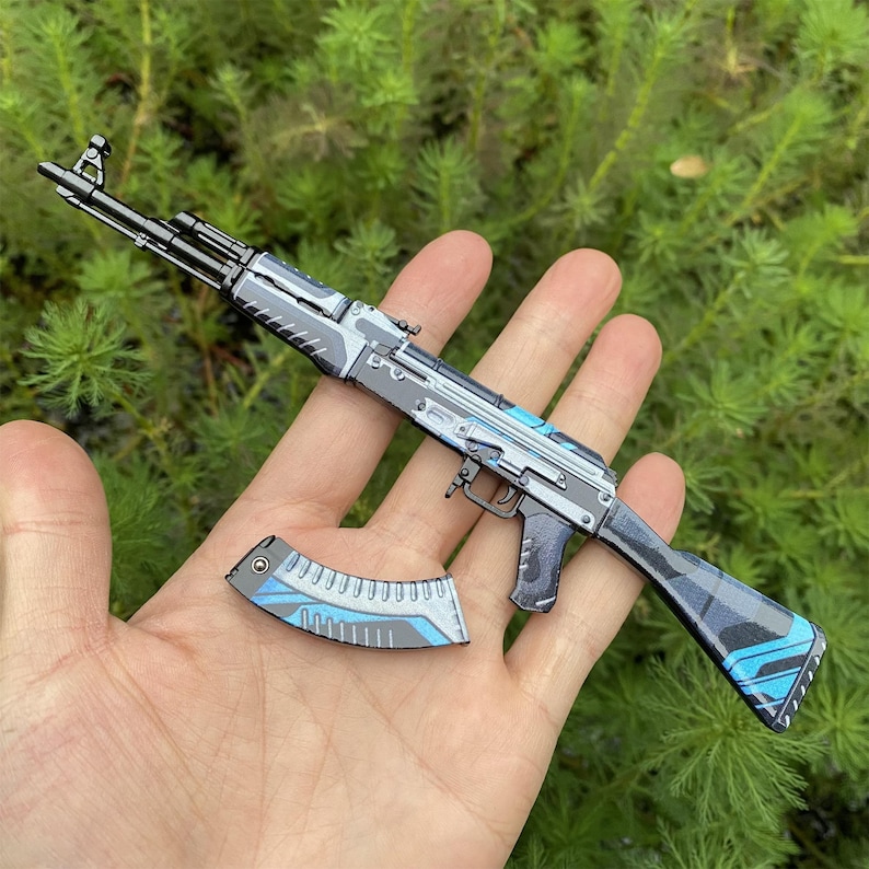 May include: A miniature AK-47 assault rifle with a detached magazine. The rifle features a black, gray, and blue color scheme with intricate detailing. The magazine is gray and blue. The background is blurred green foliage.