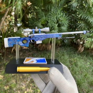 May include: A miniature sniper rifle model featuring a blue and white body with intricate swirling patterns, a silver scope, and a bipod. The model is mounted on a black rectangular stand that includes a yellow bullet and a blue and orange logo.