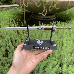 May include: A replica sword in silver and copper tones, presented on a black stand. The sword has detailed handle designs and a sharp blade. The stand is inscribed with "The Elder Scrolls" and a logo.
