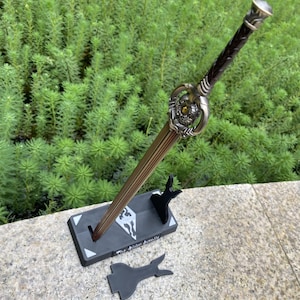 Pu&ograve; includere: Una spada decorativa con una lama in bronzo e un'elsa ornata, esposta su un supporto nero. L'elsa presenta un design dettagliato con una gemma gialla. Il supporto ha un logo e il testo "The Elder Scrolls V: Skyrim".