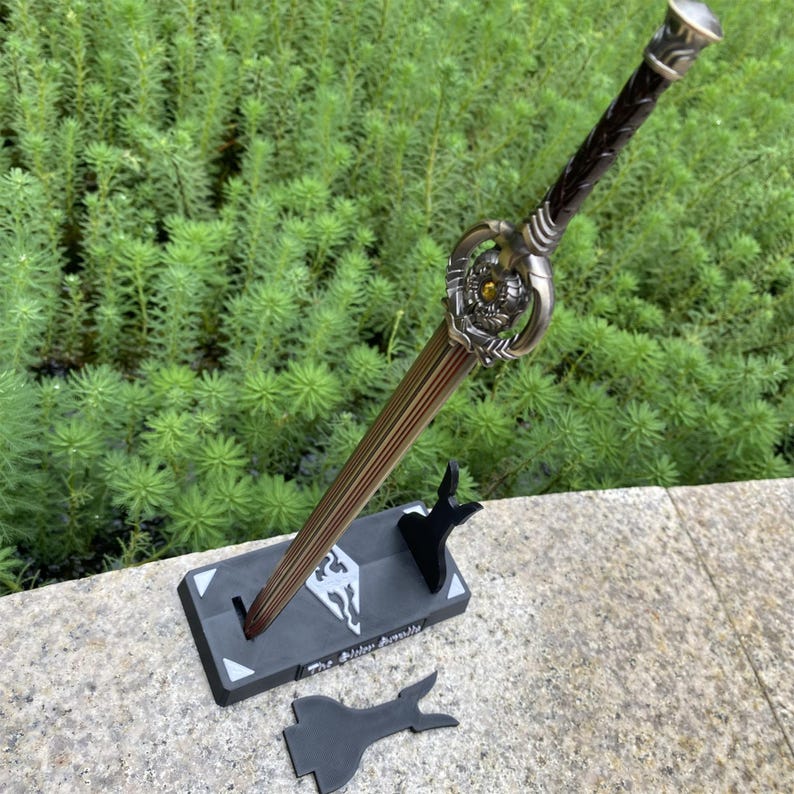 Pu&ograve; includere: Una spada decorativa con una lama in bronzo e un'elsa ornata, esposta su un supporto nero. L'elsa presenta un design dettagliato con una gemma gialla. Il supporto ha un logo e il testo "The Elder Scrolls V: Skyrim".