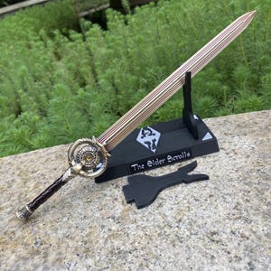 Pu&ograve; includere: Una spada decorativa con lama e impugnatura dorate, appoggiata su un supporto nero. La lama presenta un design a strisce con accenti rossi. Il supporto presenta il testo "The Elder Scrolls" e un simbolo.