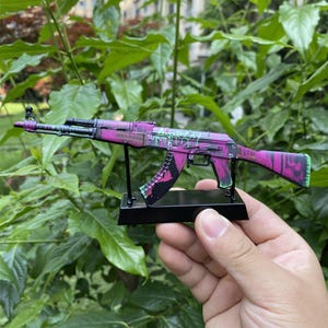 以下が含まれることがあります： 鮮やかなピンクとグリーンの落書きデザインのミニチュアAK-47ライフル。おもちゃの銃は黒いスタンドに取り付けられており、その詳細な特徴を示しています。背景は緑豊かな葉です。
