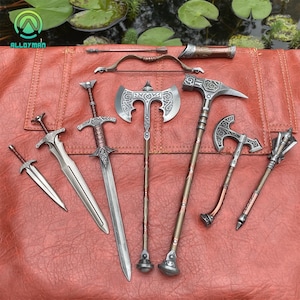 May include: A collection of miniature metal weapons, including swords, axes, a hammer, and a bow and arrow, are displayed on a textured, reddish-brown surface. The weapons feature detailed designs and are likely decorative items.