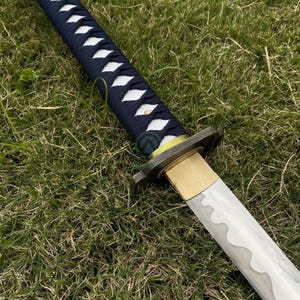 Może przedstawiać: Japoński miecz katana z granatowym uchwytem owiniętym białymi, diamentowymi akcentami. Ostrze jest srebrne z falistą krawędzią, a osłona jest złota. Miecz spoczywa na zielonej trawie.