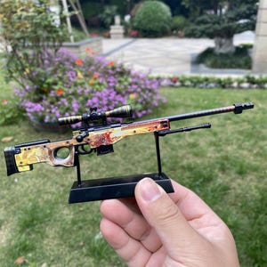 Op de afbeelding: Een gedetailleerde miniatuur sluipschuttergeweer met een vizier, gepresenteerd op een zwarte standaard. Het geweer heeft een beige en rood decoratief ontwerp. De vizier is zwart en goudkleurig. Het ontwerp is gedetailleerd en decoratief.