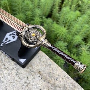 Pu&ograve; includere: Una spada replica dettagliata con un'elsa color bronzo e un'impugnatura intrecciata marrone. La lama presenta un design oro e rosso. La spada poggia su un supporto nero con un emblema bianco. Il design suggerisce un tema fantasy o videoludico.