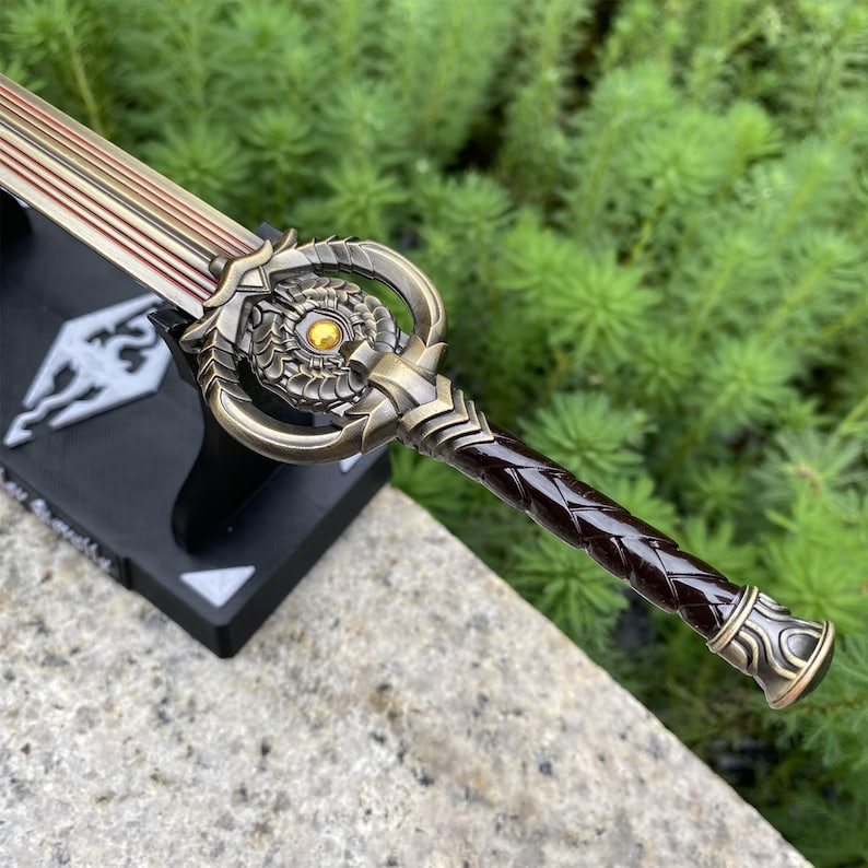 Pu&ograve; includere: Una spada replica dettagliata con un'elsa color bronzo e un'impugnatura intrecciata marrone. La lama presenta un design oro e rosso. La spada poggia su un supporto nero con un emblema bianco. Il design suggerisce un tema fantasy o videoludico.