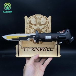 Può includere: Un coltello nero e argento con un accento giallo e un'impugnatura nera con un pulsante rosso. Il coltello è esposto in un supporto in legno con un logo Titanfall e un design a testa di robot.