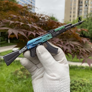 以下が含まれることがあります： 青と緑のグラデーション仕上げのミニチュアAK-47スタイルのライフル。おもちゃの銃は黒いマガジンとストックがあり、上部に数字の「20」が見えます。画像は屋外で撮影されました。