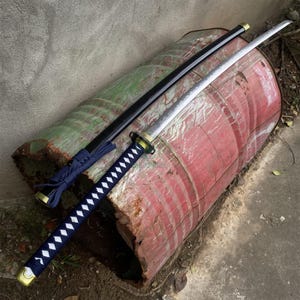Può includere: Una spada katana con una lama argentata e un fodero nero. L'impugnatura è avvolta in tessuto blu con motivi a rombi bianchi. La spada poggia su un tamburo di metallo rosso e verde usurato.